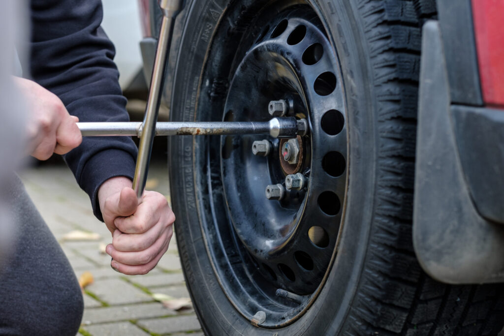 Close up of 2 hand using a tire iron to loose the lug nuts of a tire sitting on pavement outside