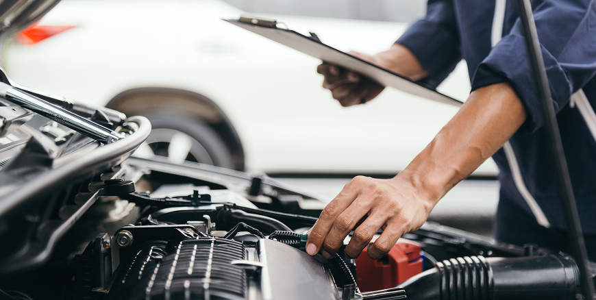 A Telle Tire mechanic holding a clipboard, checking various components of a vehicle's engine.